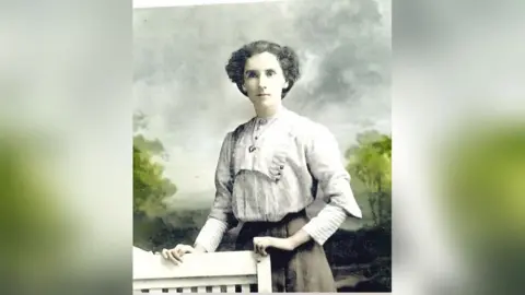 A lady stands holding onto a chair in what appears to be a very old photograph. She is wearing a blouse and skirt and is in a photography studio with a tree lined background behind her
