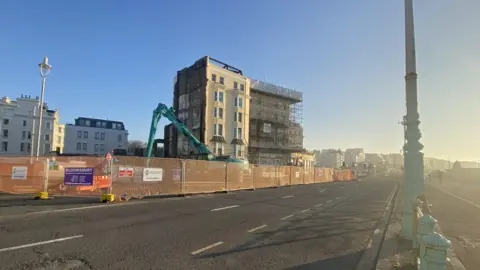 Empty three-lane road on Brighton seafront with fire damaged hotel fenced off in the background.