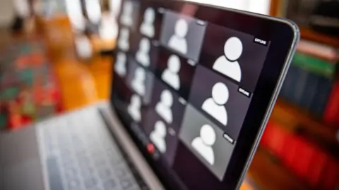 A laptop is on desk, with the rest of the room blurred. There are anonymous squares of people waiting to join an online meeting.