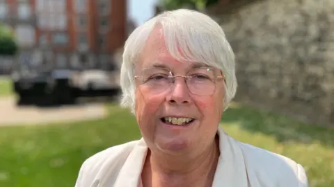Andrew Sinclair/BBC Charlotte Cain, MP for Ely and East Cambridgeshire wearing a white jacket and glasses, smiles for the camera.