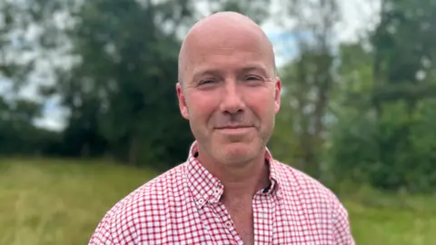 A man with a bald head and a red and white checked shirt standing in a green field