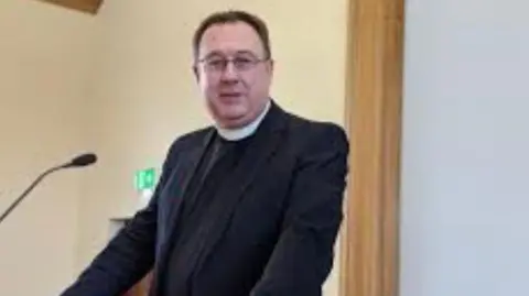 Handout A man in a dark outfit and white collar looking at the camera from a lectern. There is a microphone in front of him and a fire exit sign behind him