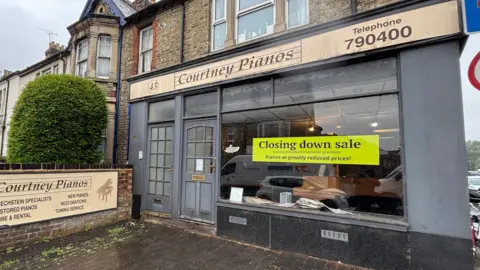 The front facade of Courtney Pianos on the Botley Road, with a large bright 'closing down' sign in its window.