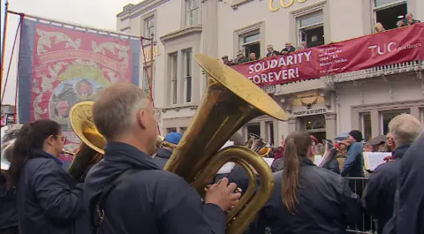 A brass band playing in front of a banner saying Solidarity Forever, hung from a hotel balcony
