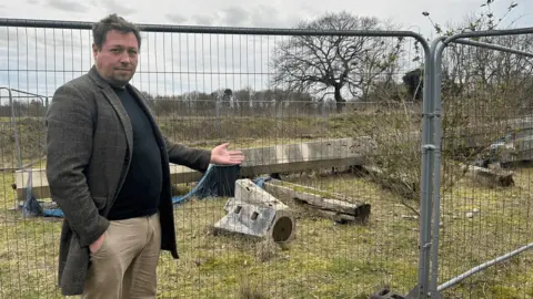 Rory Handy wearing a dark-green-brown blazer with a black shirt underneath. He is standing in front of a grey fence and pointing his hand towards it. Behind the fence is a large wooden structure dumped on the ground. There are trees in the background. 