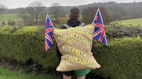 A man with dark hair stands with his back to the camera with a sack on his back with the words weather, finances, exhaustion, loneliness and regulations written across it.