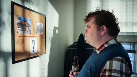 BBC/Two Brothers/Matt Squire Ethan Lawrence as Ben in Boat Story. Ben is pictured off duty, holding a beer and looking at a cork board with two pictures connected by a red thread with a question mark printed underneath. Ben is a man in his 30s with curly brown hair and wears a blue vest over a red and white plaid shirt 