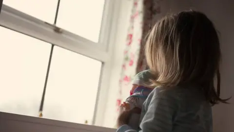 A girl with light brown hair and holding a doll looks out of a window. She is wearing a striped T-shirt.