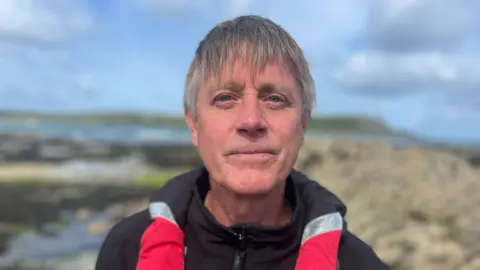A man looking directly at the camera, he is wearing a black zip up jacket with red and silver. Behind him the background is blurred. It is a rocky terrain and there is the sea in the background. 