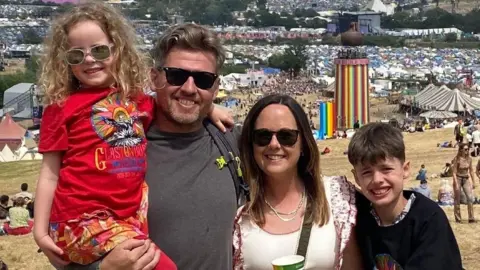 Kelly, her husband and children at Glastonbury Festival. Her husband is second from left and wearing a grey t-shirt and shades, holding their young daughter in his arms, she is wearing a red Glastonbury t shirt and red leggings. Kelly is wearing a white t shirt, sunglasses and holding a cup of drink and their son is wearing a dark top. The whole family is smiling at the photographer with some bright rainbow tents in the background.