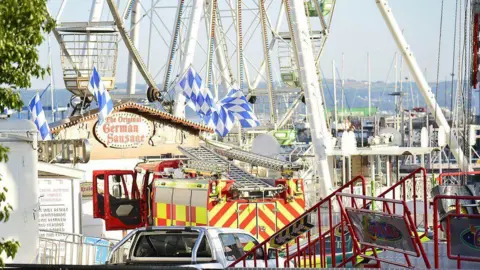 Pacemaker A fire engine is parked in a fun fair beside rides. The doors are open. No one is about. The grounds appear to be empty.