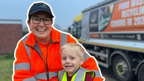 Binlady smiles to camera with her arm around a little boy with blonde hair. She is wearing a bright fluorescent orange hi-vis jacket, a black cap and glasses. The boy is smiling wearing a yellow hi-vis jacket. A bin lorry can be seen in the background parked outside a residential property with a front garden.