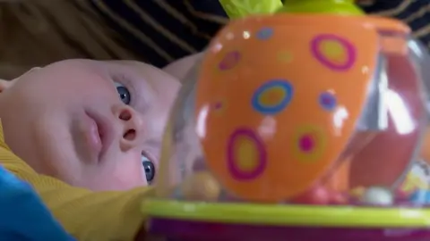 BBC The image shows a baby with large blue eyes, lying on the floor looking at a see-through circular plastic toy, with small coloured balls inside it. The little boy is wearing a mustard-coloured top.