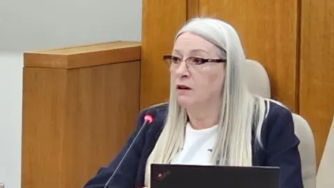 Kay Mason Billig is sitting at a council meeting. She has a navy jacket on top of a white top.