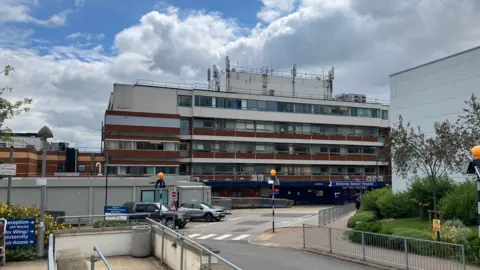 Sam Read/BBC A photo of Kettering General Hospital, a multi-storey building which looks run down. There is construction site in the foreground and a number of cars parked besides a zebra crossing. 