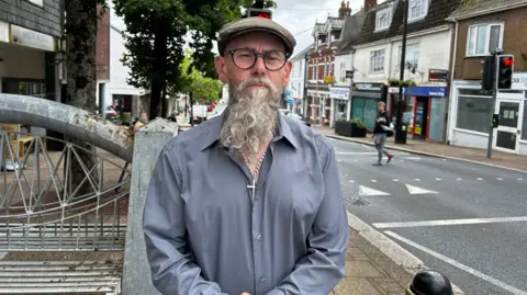 A man with a long grey beard, wearing a blue shirt and a brown cap and glasses. He is looking directly at the camera. Behind him to the right is a road and traffic lights. To the left of him is the high street and a metal bench. 