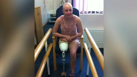 Cancer Reasearch UK handout Liv Shaw in a medical treatment room after she had part of her leg amputated at the age of 14. She is sitting in a chair and is wearing a pale pink sleeveless dress. She is smiling.