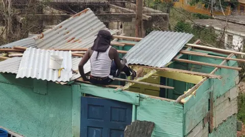 Family Handout A man is sitting on the broken roof of his home trying to repair it. The house is made of wooden slabs and metal.
