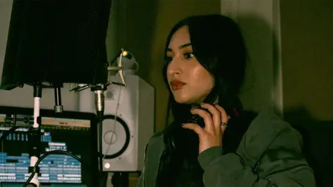 @Casa.Lente Khushi Kaur, a female singer in a music studio, looking into a microphone while holding a black pair of headphones around her neck. She is wearing a green jacket, with a computer screen and white speakers in the background.