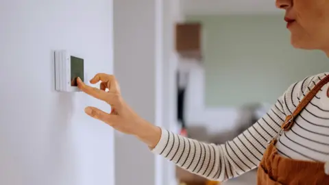 A woman is pictured adjusting the heating using a wall mounted electronic panel. She is dressed in a blue and white stripe top, with copper coloured dungarees. 