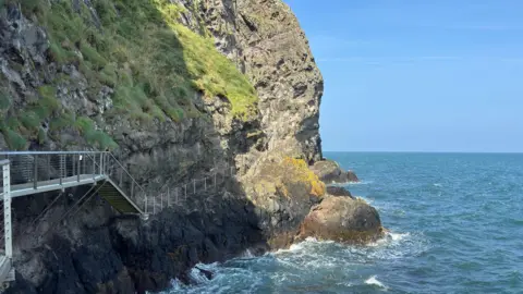 Getty Images The Gobbins cliff face is grey and brown with green grass growing on parts of it. There is a silver path and steps running along the cliff face. Water is crashing up against the cliff face, it is blue and white. The sky is blue in the background. 
