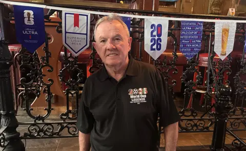 Pratiksha Ghildial / BBC Enda Keenan stands in front of flags for the 2026 world cup