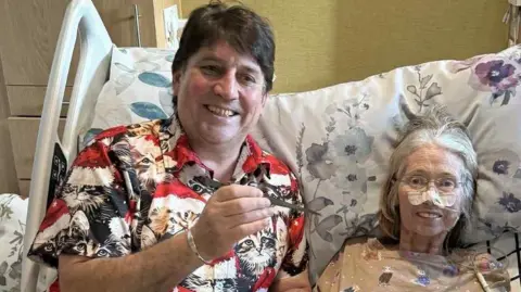 John Owen John and Rowena Owen sit in a large hospital type bed. Rowena has a tube going into her nose, held in place by micropore tape. She is wearing a top with cats on while John wears a shirt with cats on. The are both smiling. Rowena is looking into camera but John is looking slightly off camera.