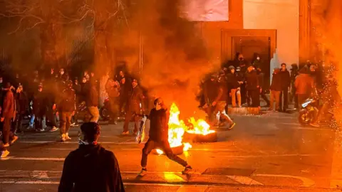 Getty Images Protesters dressed in black gather as a fire burns on the streets of Tehran, Iran.