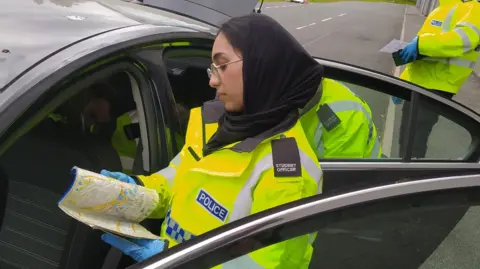 West Yorkshire Police A police officer wearing a high-visibility jacket and blue gloves standing beside an open car door. The officer is holding a folded map. Another officer in similar attire is visible in the background, holding a clipboard or document.