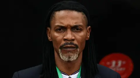 A close-up headshot of Rigobert Song looking sternly into the camera while wearing a black jacket, white shirt and with green lanyard around his neck