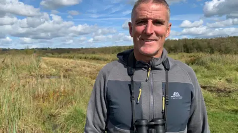 Wildlife naturalist Iolo Williams standing on Crymlyn Bog, wearing a waterproof coat and a pair of binoculars around his neck