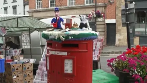 A red post box is covered in knitted yarn bombs including Postman Pat and a couple of animals