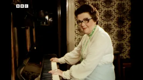 A screenshot from the BBC Look North clip that shows Peggy Fullerton playing the piano while looking into the camera. She's got short, curly brown hair and large frame glasses. She's wearing a white shirt with green beaded necklace. Behind her is a dark brown door and patterned wallpaper.