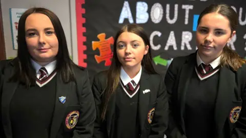 Pictured from left to right is: Cassie, Taigan and Eimear. Cassie has shoulder length black hair, Taigan has long brown hair and Eimear has a dark ponytail. All of the girls are wearing a black blazer which consists of a black blazer, black v-neck jumper, striped burgundy and white tie, and a white shirt.