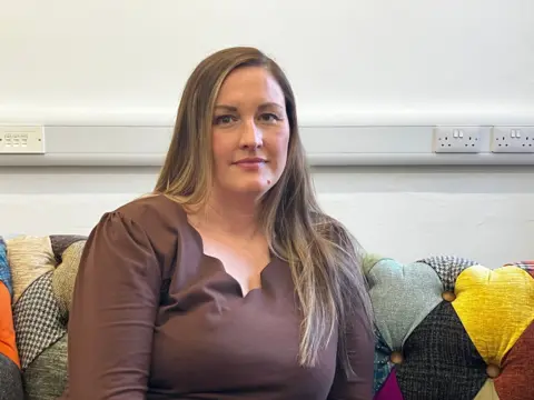 BBC News A woman sits on a bright diamond-patterned sofa. She has long brown hair and wears a brown scalloped, v-neck top