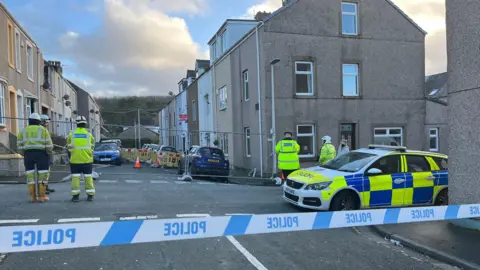 Officers in high-vis jackets stand on a street, which is partly fenced off. A police car and police tape are in the foreground.