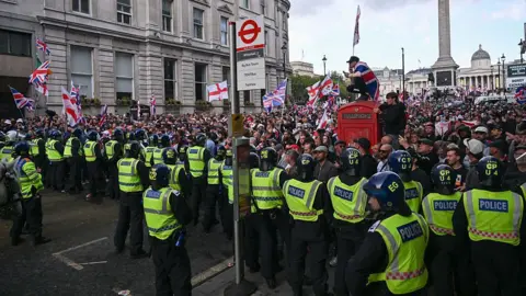 A line of police in high-visibility jackets and helmets faces a crowd waving Union Jacks and St George's Cross flags. A person draped in a flag sits atop a red telephone box near a Whitehall bus stop, with Nelson's Column in the background.