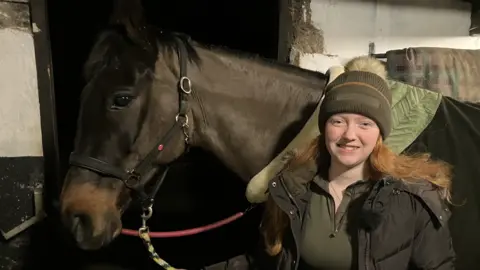 BBC News Samantha and horse Ozzy standing in a stable, Samantha smiles at the camera. Ozzy stands behind her, a black horse with a green saddle. 