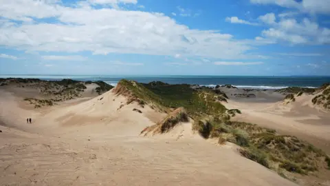 Sand dunes with a couple of people walking in the distance and the blue sea and sky in the background.