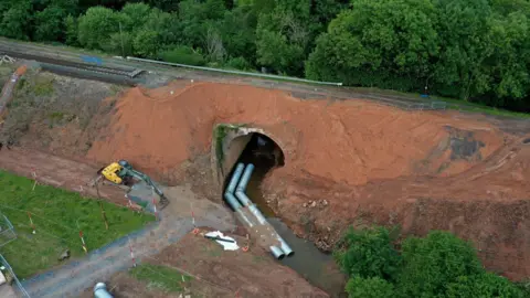 A aerial view of a railway line on top of a red earth embankment with a tunnel running through it and long metal pipes running through the tunnel. There is also a yellow mechanical digger to the left of the tunnel