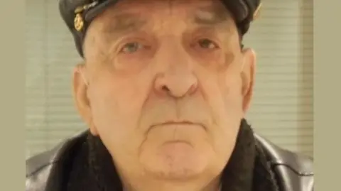 Police mugshot of a man wearing a cap with badges on the side and jacket with a furry collar. He is looking straight at the camera.