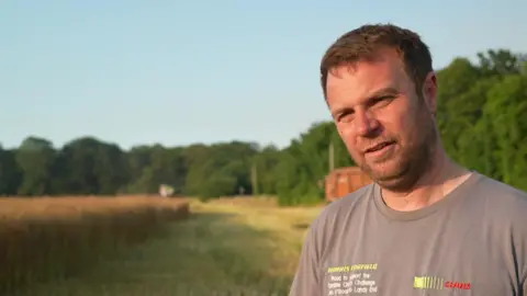 Farmer Olly Harrison with short brown hair and stubble wearing a beige t-shirt. He is standing in a field with trees behind him on a sunny day.