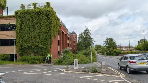 Jacob Ottaway/BBC A view of a multi-storey car park from a slight distance. A uniformed officer stands in front of the corner of the building. A silver car drives past on the road running parallel. 