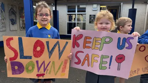 Two young girls stand outside a primary school. They are both dressed in a school uniform of a blue jumper and a yellow polo shirt underneath. The girl on the left holds a paper sign with the words 'Slow Down' written in colorful lettering. Beside her another girl holds a similar hand drawn sign with the words 'keep us safe" in bright bubble letters.