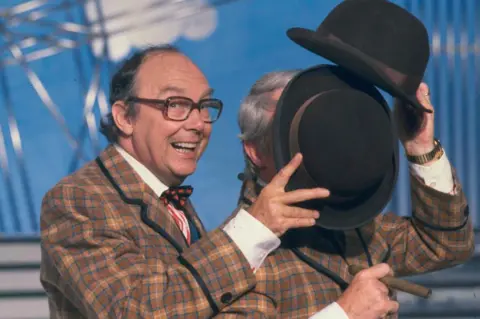 Getty Images Eric Morecambe is grinning as he holds his hat to block Ernie Wise's face during a dance routine