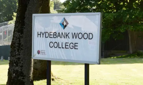 An entrance sign saying 'Hydebank Wood College' - the words are in black typeface on a white background. The sign is outside on a grass verge and trees can be seen in the background. 