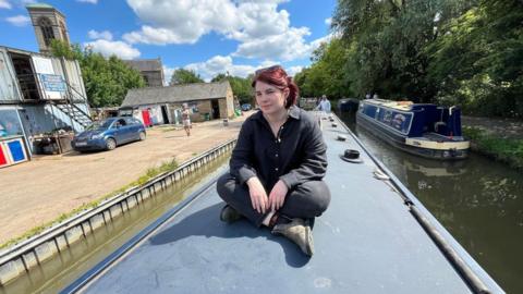BBC Reporter Clodagh Stenson sits on the roof of a narrowboat on the Oxford Canal. She is wearing black trousers and a black shirt with sunglasses on top of her head. She has short red hair. There is a second blue narrowboat in the water alongside. One bank has green trees overhanging the water. The other bank has a gravel yard with a blue car parked next to a two-storey industrial building.