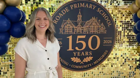 James Grant/BBC A woman in a white dress stands in front of a gold sequinned wall with blue balloons and a large blue circle with "Roade Primary School, 150 years" written in orange/ gold on it. 