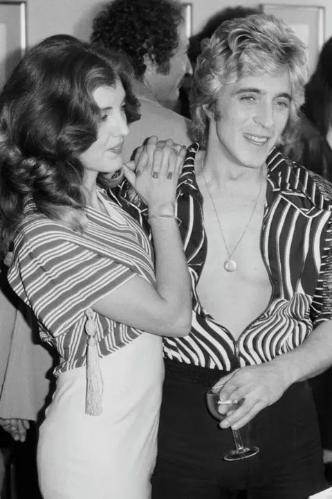 Michael Putland/Getty Images A black and white photo of a woman, Suzi Ronson, with dark wavy-styled hair just past her shoulders wearing a stripy top with wide arms and a white pencil skirt. She has her hand intertwined and resting on a man's shoulders who is stood next to her. The man, Mick Ronson, is wearing a stripy unbuttoned shirt with a necklace with a circular medallion. He has a wine glass in his hand and is smiling to the side with thick blond hair.