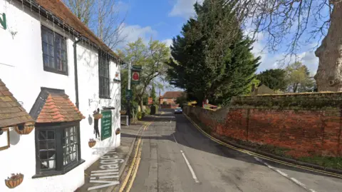 Google Village Road on streetview, showing a white pub, a narrow road and a red brick wall